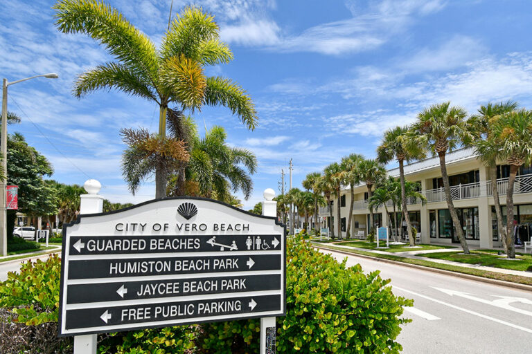Downtown Vero Beach sign with directions to local beaches and public parking, surrounded by palm trees and shopping plaza.