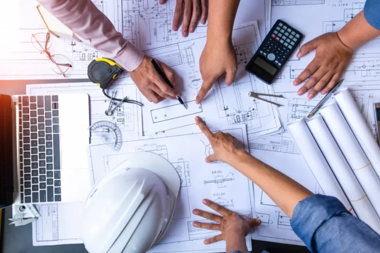 A team of professionals collaborating on home blueprints, showing hands pointing to construction plans on a table with a hard hat, calculator, and laptop nearby.